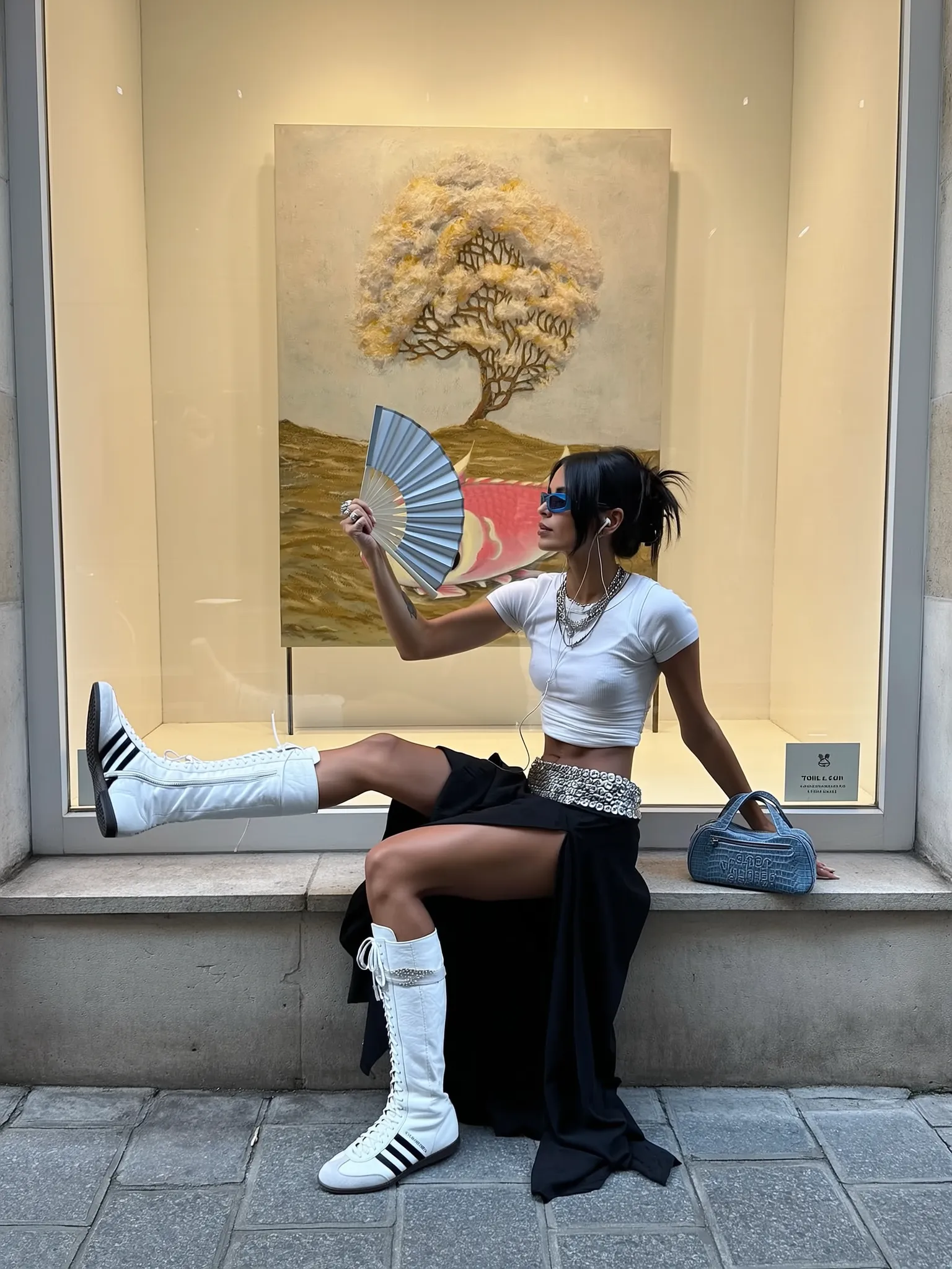 A high-contrast editorial-style street photograph of a woman sitting on the stone ledge of a gallery window in a narrow urban alleyway. Behind her, through the glass, a textured painting of a distorted tree and pink fish hovers on a pale cream wall, adding an abstract artistic backdrop. The woman is positioned in semi-profile, her legs extended across the stone surface and the patterned concrete sidewalk. She wears a fitted white ribbed crop tank that clings tightly to her torso, revealing a toned midriff. Her bottoms are dramatic — a black asymmetrical skirt with high slits that exposes her legs and flows down in loose ruffles. Around her waist is a thick, silver-buckled statement belt layered over the skirt. On her feet: striking white high-top Adidas boxing boots with black stripes and tight lacing, contrasting against the soft femininity of her outfit. One foot rests elevated, toe-pointed outward, giving her posture a defiant ease. She holds a pale blue folding hand fan mid-motion in her left hand, fingers adorned with chunky silver rings, her wrist relaxed. Her right arm is behind her, balancing her on the ledge, next to a textured mini denim handbag with embossed white monogram text. Her face is framed by two loose strands of jet-black hair while the rest is pulled back into a messy ponytail. She wears rectangular blue-tinted sunglasses, and a pair of earphones are subtly visible. Multiple chain necklaces adorn her neck. The lighting is natural, soft yet directional — late afternoon tones, casting faint shadows. The whole scene captures a mix of hyper-modern streetwear and cool indifference against an artful urban backdrop.