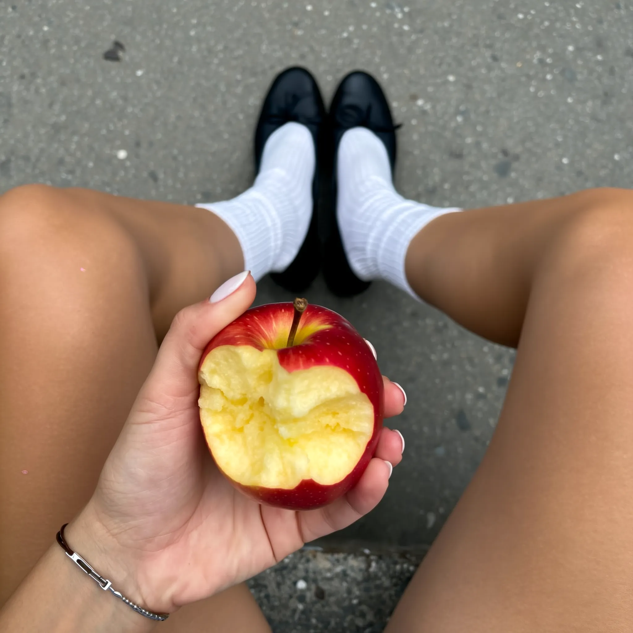 SHOT: • Composition: Top-down close-up view, apple centered in hand, legs spread in background • Camera Settings: Natural light, moderate depth, standard lens around 35mm • Film Grain: Smooth, natural digital clarity LENS EFFECTS: • Optics: Sharp focus on bitten apple and hand, background slightly blurred • Artifacts: None • Depth of Field: Shallow, emphasizing apple and hand with legs and ground softly out of focus SUBJECT: • Description: Hand holding red and yellow bitten apple • Wardrobe: White ribbed calf socks, black ballet flats • Grooming: Clean, natural hand skin tone, no accessories SCENE: • Location: Outdoors on a textured concrete or stone surface near asphalt • Time of Day: Daytime with soft natural lighting • Environment: Urban pavement with visible speckles and dirt VISUAL DETAILS: • Action: Hand holding apple directly towards viewer • Props: Bitten apple, socks, ballet flats • Physics: Gravity apparent in seated posture and held apple CINEMATOGRAPHY: • Lighting: Soft diffuse daylight, evenly lit • Tone: Casual, relaxed • Color Palette: Muted neutrals with bright red and yellow of apple, white socks, black shoes TEXT ELEMENTS: • Visible Text: None • Typography: None • Placement: None STYLE: • Visual Aesthetic: Minimalist, candid snapshot