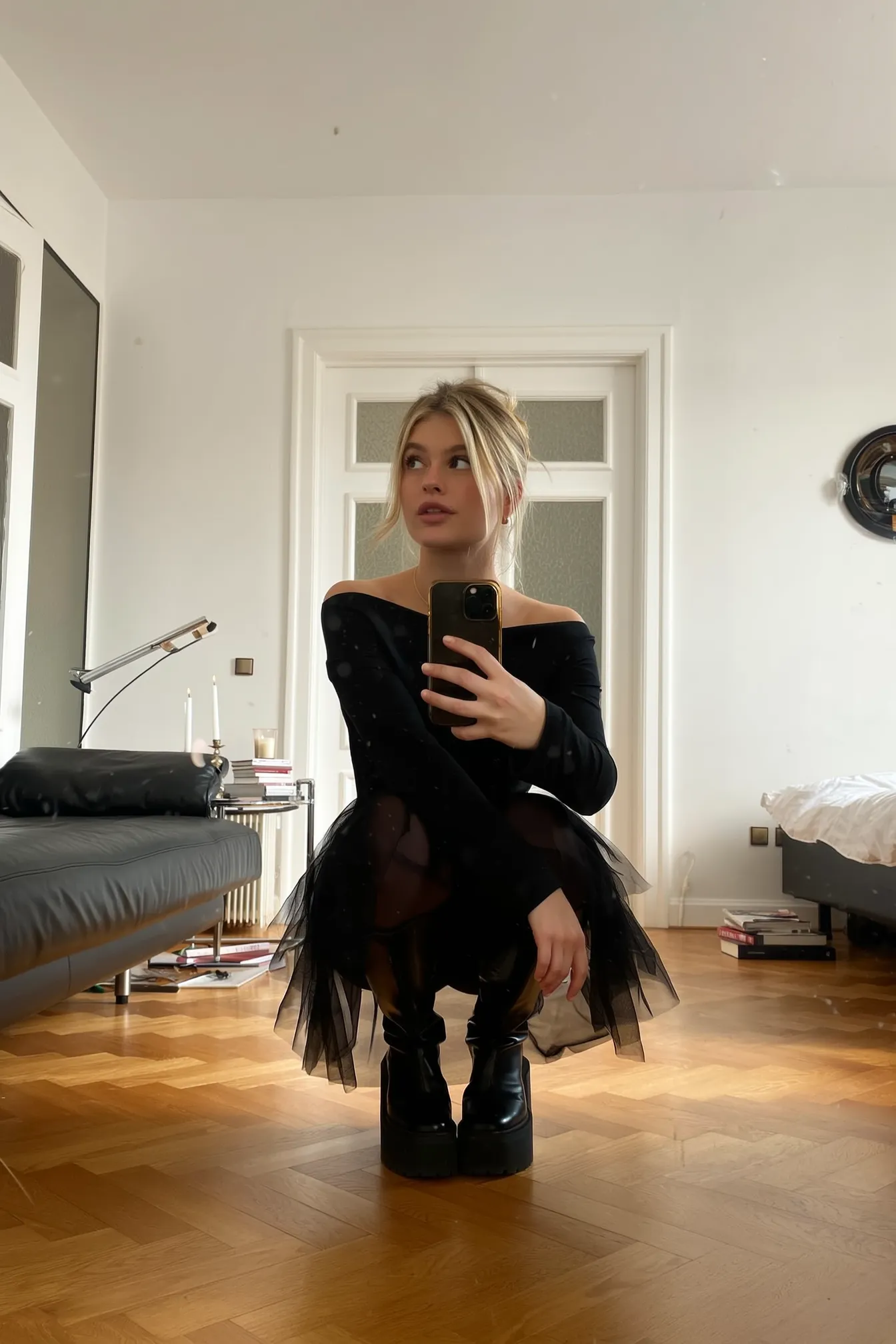 SHOT: • Composition: Centered full-body shot, low angle, person squatting in middle foreground with room elements in background • Camera Settings: Neutral exposure, natural indoor lighting, smartphone camera • Film Grain: None, clear digital image LENS EFFECTS: • Optics: Sharp focus on subject, soft background detail • Artifacts: None • Depth of Field: Moderate, subject in clear focus, background slightly blurred SUBJECT: • Description: Young woman with blonde hair tied back, looking slightly to left, holding phone for mirror selfie • Wardrobe: Black sheer layered dress with off-shoulder style, black chunky platform boots • Grooming: Natural makeup, hair loosely tied up with some strands falling SCENE: • Location: Interior of a modern, minimally furnished living room • Time of Day: Daytime, natural light coming from window outside frame • Environment: Wooden parquet flooring, white walls, white double doors with frosted glass panels in background VISUAL DETAILS: • Action: Squatting pose, holding smartphone at chest level with right hand, looking off-camera • Props: Black leather couch on left, small chrome side table with candles, desk lamp, stacked books, unmade bed on right • Physics: Natural daylight casting soft shadows CINEMATOGRAPHY: • Lighting: Soft, natural light, even illumination • Tone: Warm, casual, slightly candid feel • Color Palette: Neutral tones - whites, blacks, browns, warm wood floor hues TEXT ELEMENTS: • Visible Text: None • Typography: None • Placement: None STYLE: • Visual Aesthetic: Contemporary casual indoor portrait with slight artistic moodiness