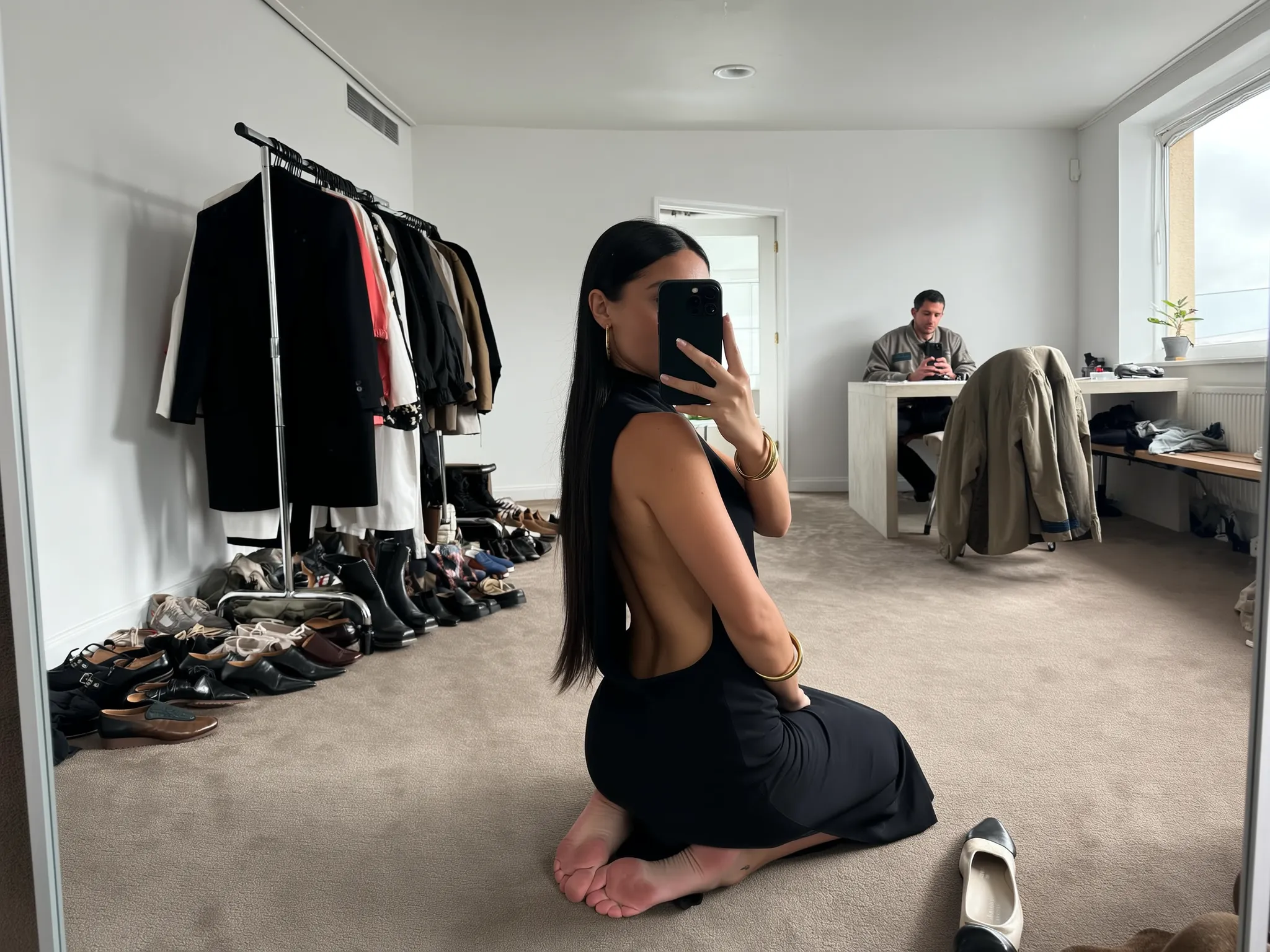 SHOT: • Composition: Medium full shot, eye-level angle, subject centered back-facing, mirror selfie style • Camera Settings: Auto white balance, ISO around 400, moderate ambient indoor light • Film Grain: None, clean digital capture LENS EFFECTS: • Optics: Sharp focus on subject and immediate surroundings, slight depth blur in background • Artifacts: None • Depth of Field: Moderate, clear subject with soft background SUBJECT: • Description: Young woman kneeling on carpet, facing away, long dark straight hair, holding smartphone covering face • Wardrobe: Sleek black sleeveless dress with large side cutouts exposing side torso and back, minimal jewelry gold bangles on wrist • Grooming: Hair straight and well-groomed, natural skin appearance SCENE: • Location: Spacious dressing room or backstage area • Time of Day: Daylight, natural light filtering through window • Environment: Casual, with clothes rack filled with various garments, boots and shoes scattered nearby, man seated at desk in background wearing casual jacket VISUAL DETAILS: • Action: Subject taking selfie with phone, barefoot with feet tucked under, one shoe near her • Props: Phone with triple camera lenses, clothing rack with assorted clothes, chair with jacket draped, various shoes on floor • Physics: Natural indoor light casting soft shadows CINEMATOGRAPHY: • Lighting: Natural diffused daylight combined with soft artificial room lighting • Tone: Neutral, relaxed, candid • Color Palette: Muted tones, black, beige carpet, earth tones in clothing and room TEXT ELEMENTS: • Visible Text: None • Typography: None • Placement: None STYLE: • Visual Aesthetic: Contemporary casual fashion photography, candid, minimalistic interior