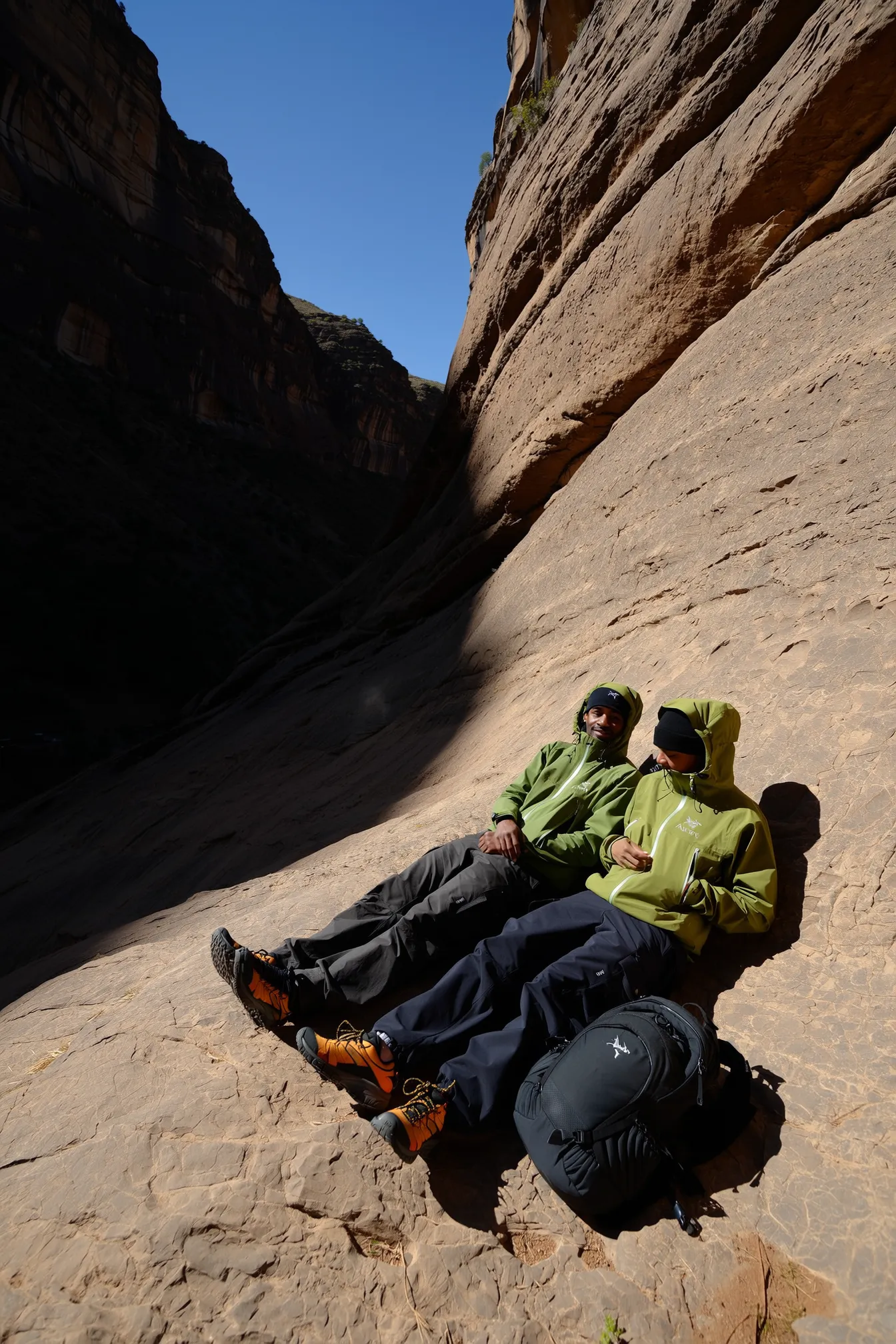 A casual, authentically candid photo spontaneously captured with an iPhone, featuring two individuals reclining comfortably against a giant limestone rock wall inside a canyon. They sit within a patch of bright sunlight that creates a natural spotlight on the textured stone surface. Both wear olive green hooded Arc'teryx jackets with adjustable cords, paired with black loose-fitting cargo pants. Their footwear consists of orange-accented technical sneakers, complemented by black beanies. Durable black backpacks rest naturally on the ground nearby. Their relaxed postures and natural body language convey genuine, casual outdoor intimacy. Surrounding cliffs are dark and textured, forming strong contrast with the warm sunlit area. The scene is illuminated by cinematic natural light with softly diffused daylight enhancing subtle fabric textures and rugged rock details. The minimalist composition frames the subjects from afar, highlighting their small scale against the vast canyon landscape, with a slightly tilted angle and film photography aesthetic. This reinforces authentic, trendy gorpcore style and believable mountainous outdoor realism.